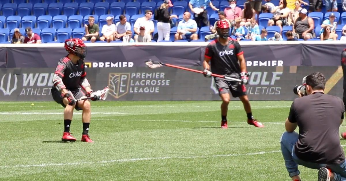 Two Defenders split the field during a Premier Lacrosse League match. Just in the frame is a photographer snapping a photo.